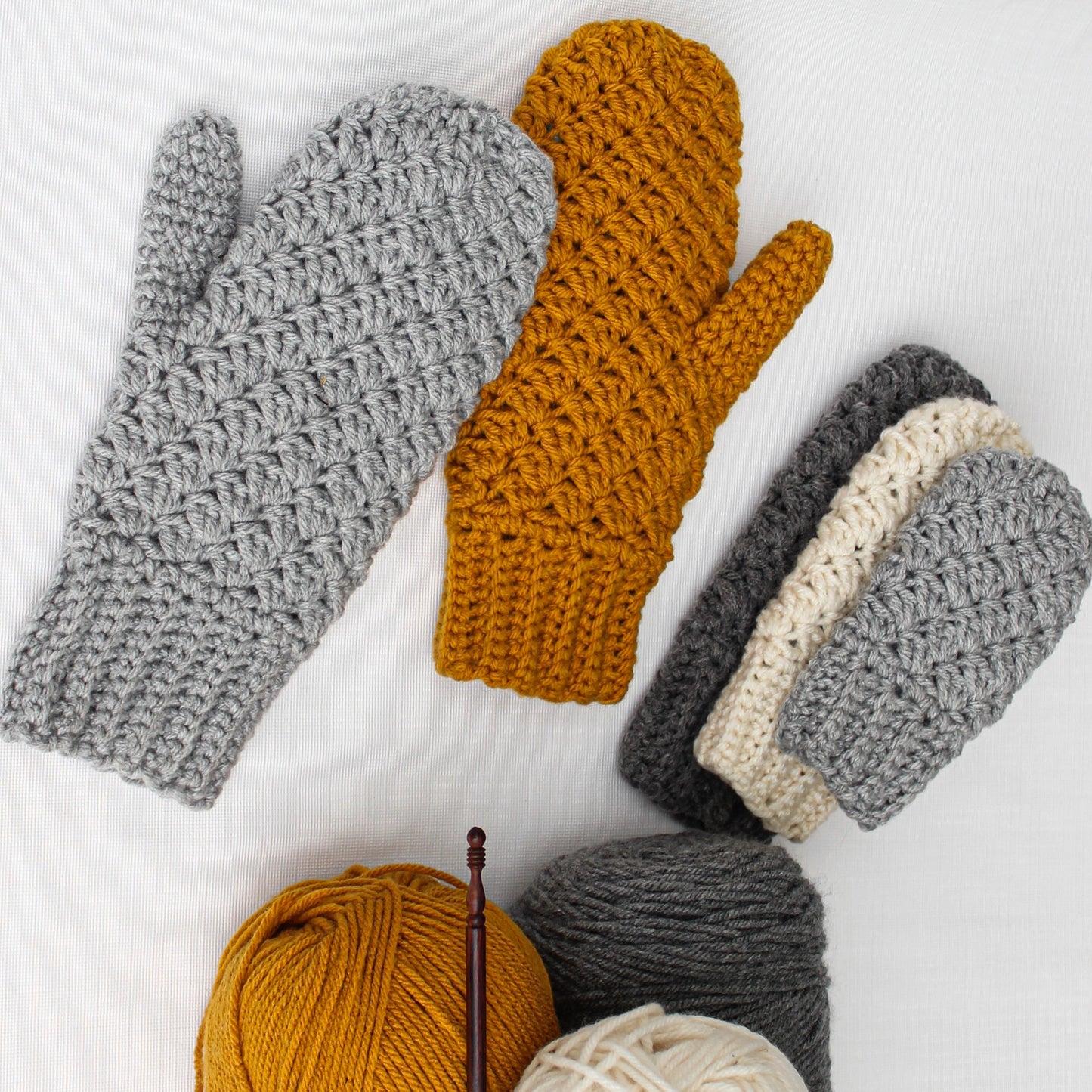 May include: A set of crocheted mittens in gray, white, and mustard yellow. The mittens are arranged on a white surface with balls of yarn in the same colors.