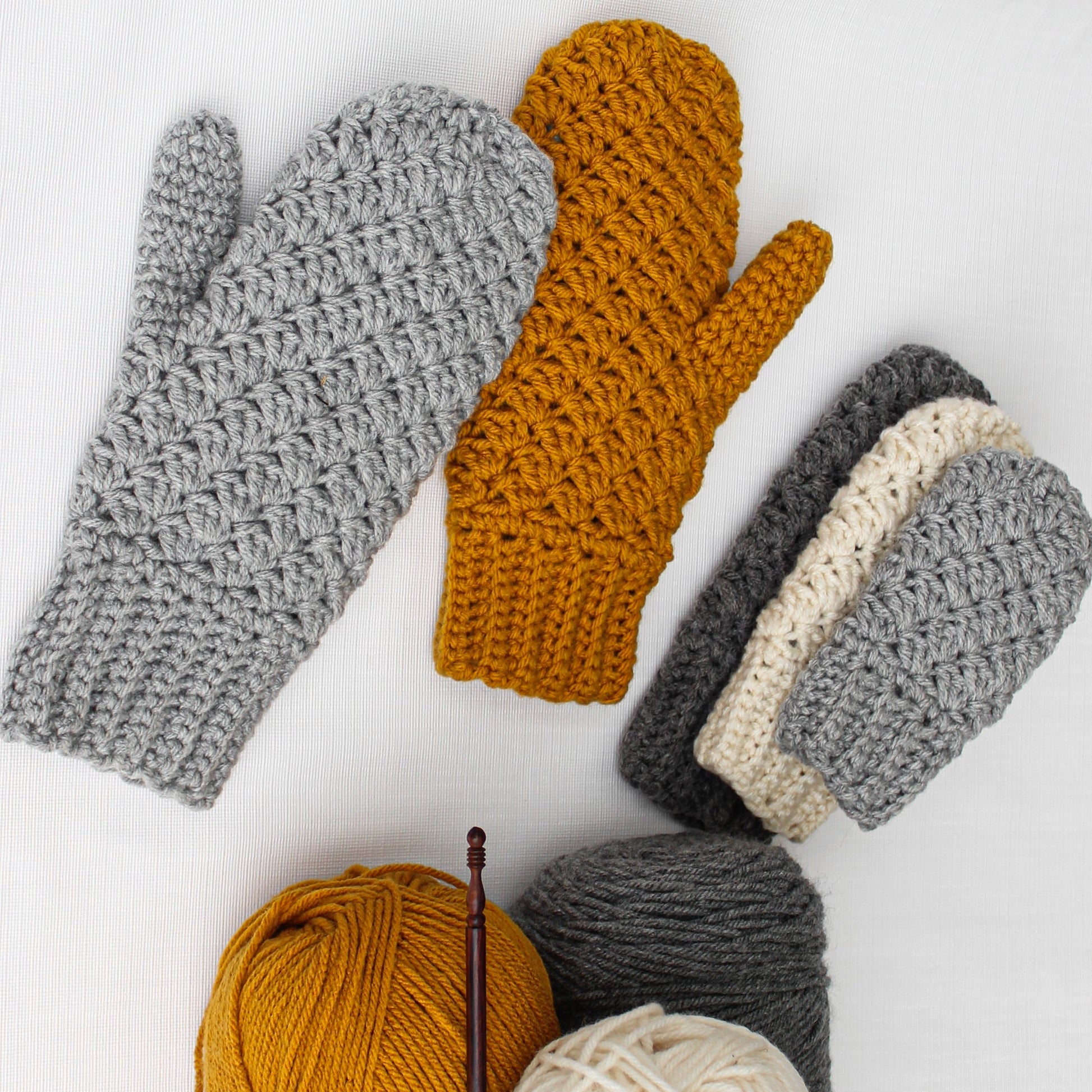 May include: A set of crocheted mittens in gray, white, and mustard yellow. The mittens are arranged on a white surface with balls of yarn in the same colors.