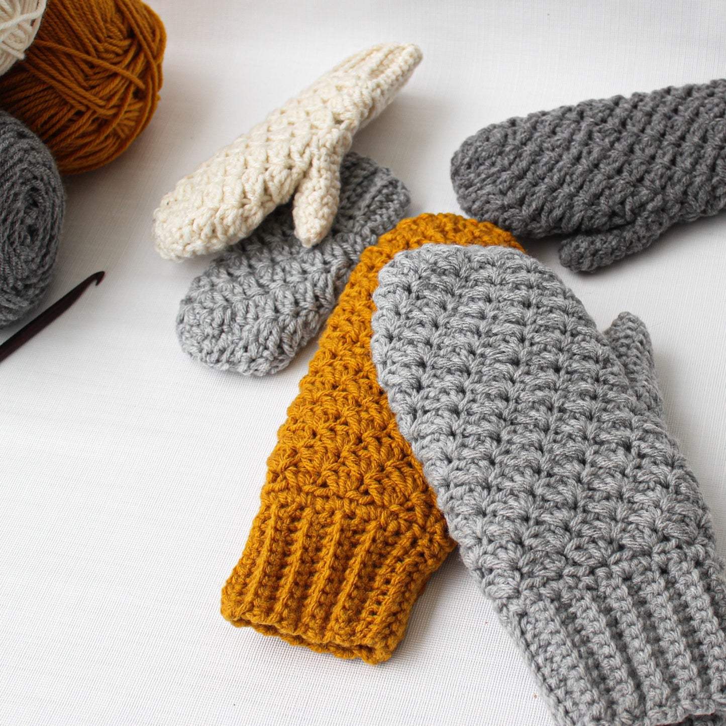 May include: Three pairs of crocheted mittens in different colors: gray, white, and mustard yellow. The mittens are made with a textured stitch and have a simple, classic design.