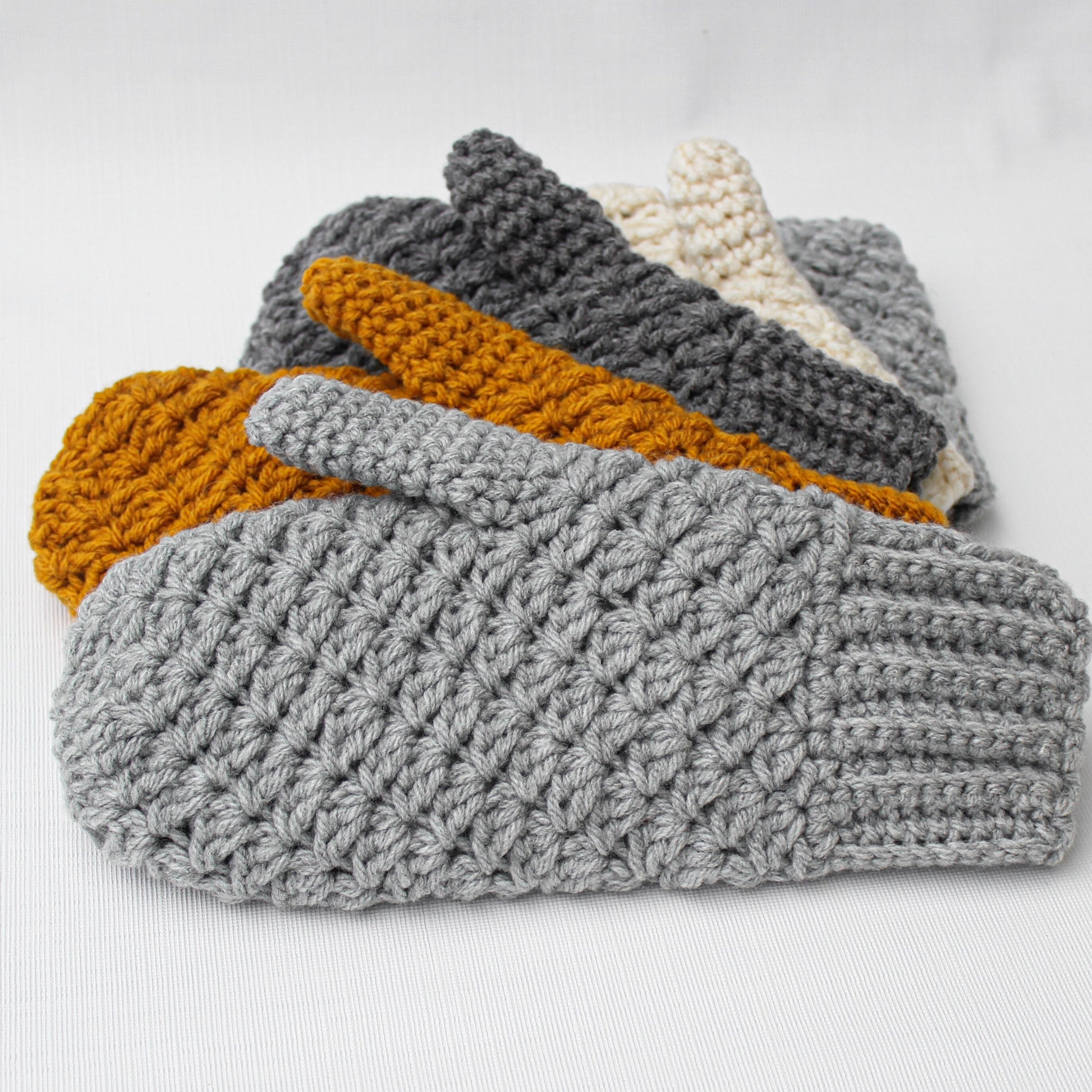 May include: A set of five crocheted mittens in various colors, including gray, mustard yellow, and cream. The mittens are stacked on top of each other, with the gray mitten on the bottom.