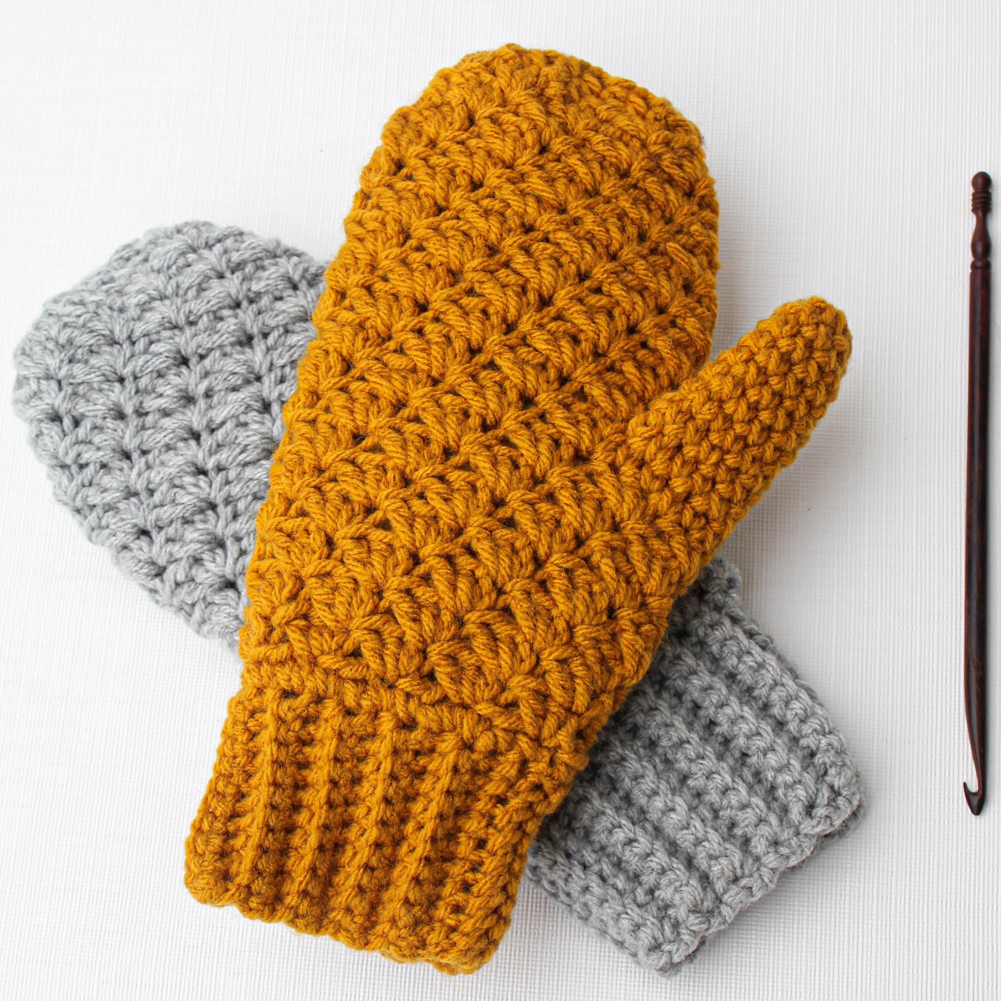 May include: A pair of crocheted mittens, one in mustard yellow and one in gray. The mittens are made with a textured stitch and have a ribbed cuff.