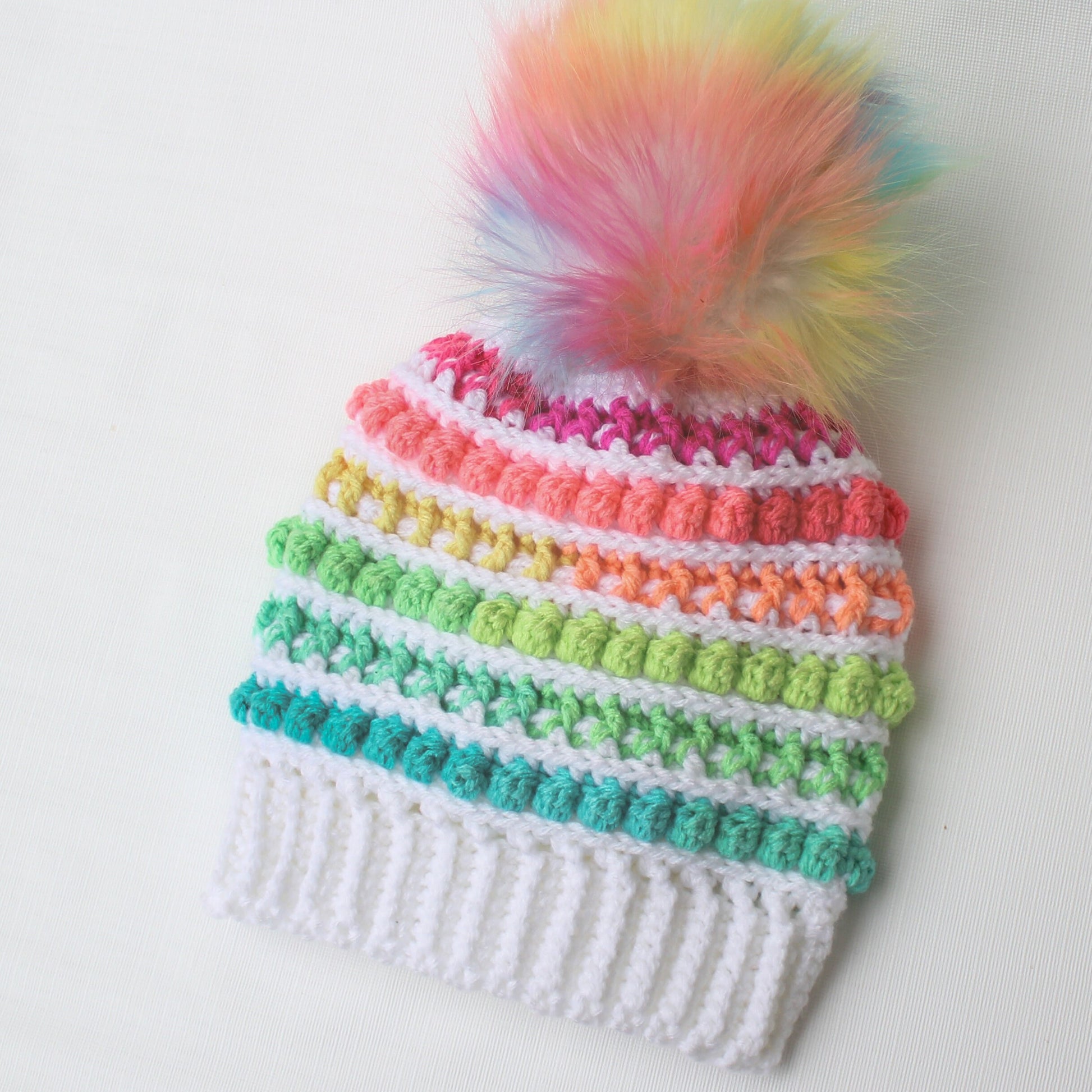 May include: A white crochet hat with a rainbow stripe pattern and a fluffy rainbow pom pom on top.