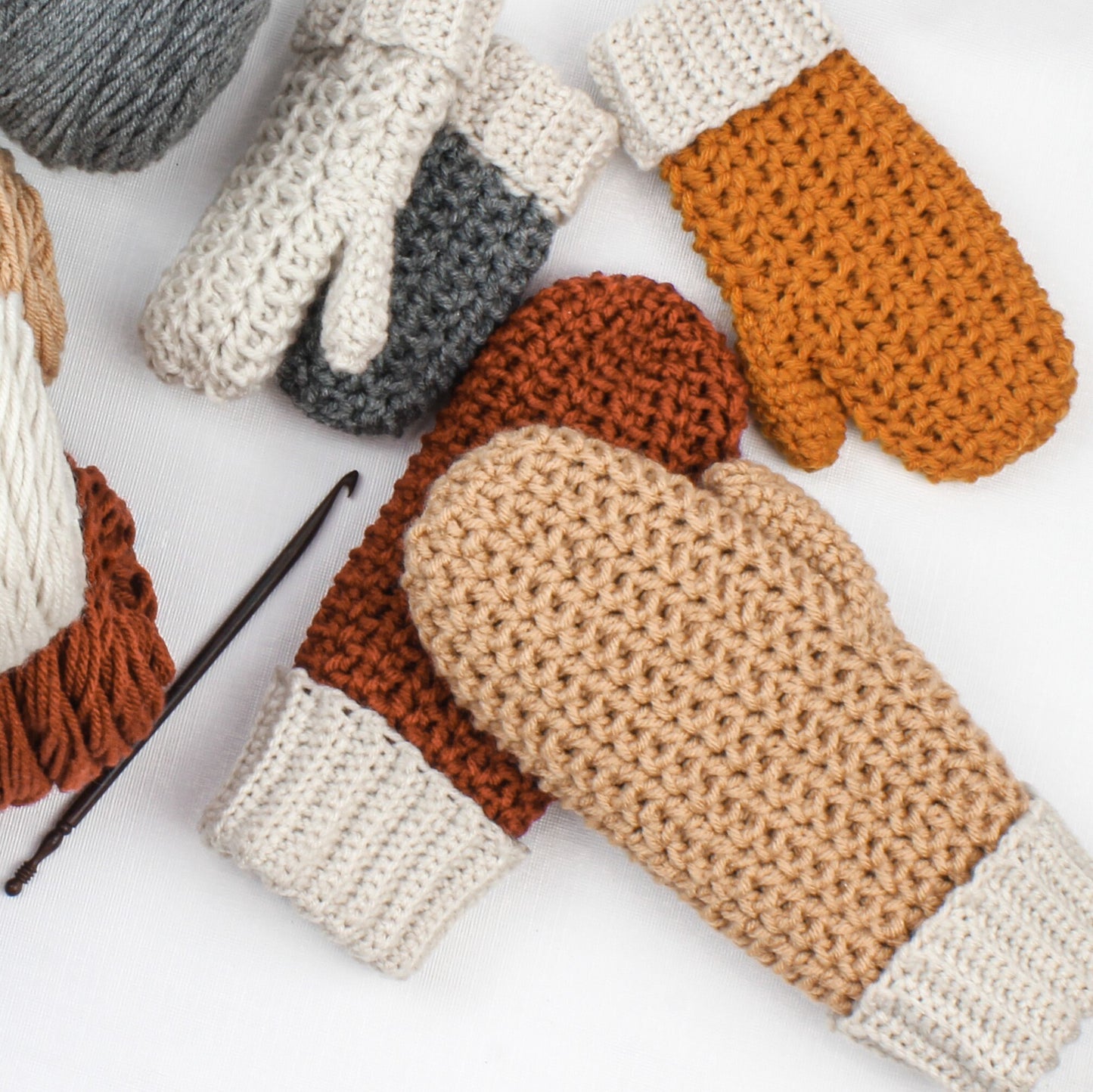 May include: Five crocheted mittens in various colors, including beige, brown, and orange. The mittens are made with a textured stitch and have a white cuff.