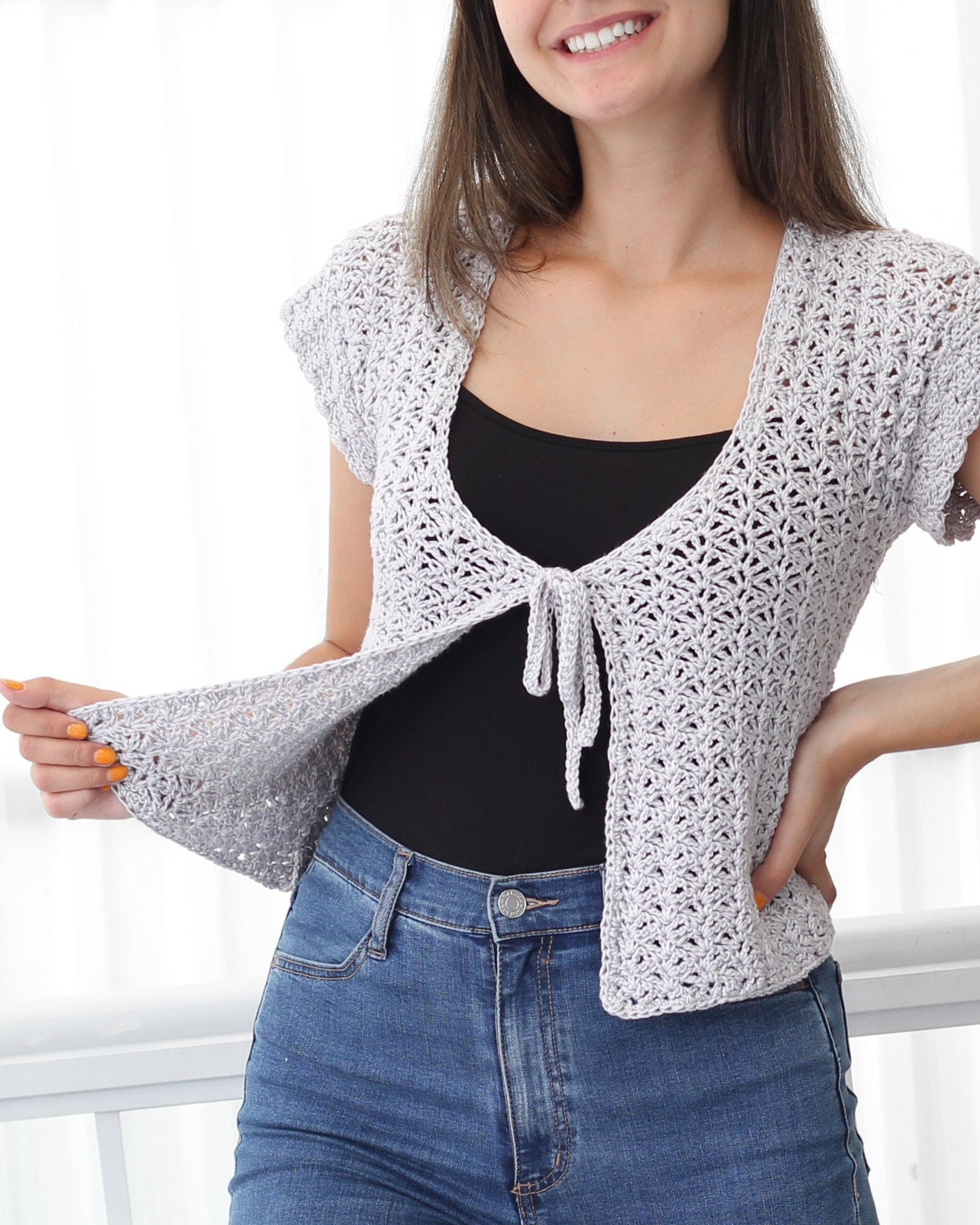 May include: A light gray crochet top with a tie front and short sleeves. The top is worn over a black tank top and blue jeans.
