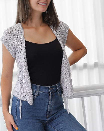 May include: A light gray crochet cardigan with a tie front and short sleeves. The cardigan is worn over a black tank top and blue jeans.