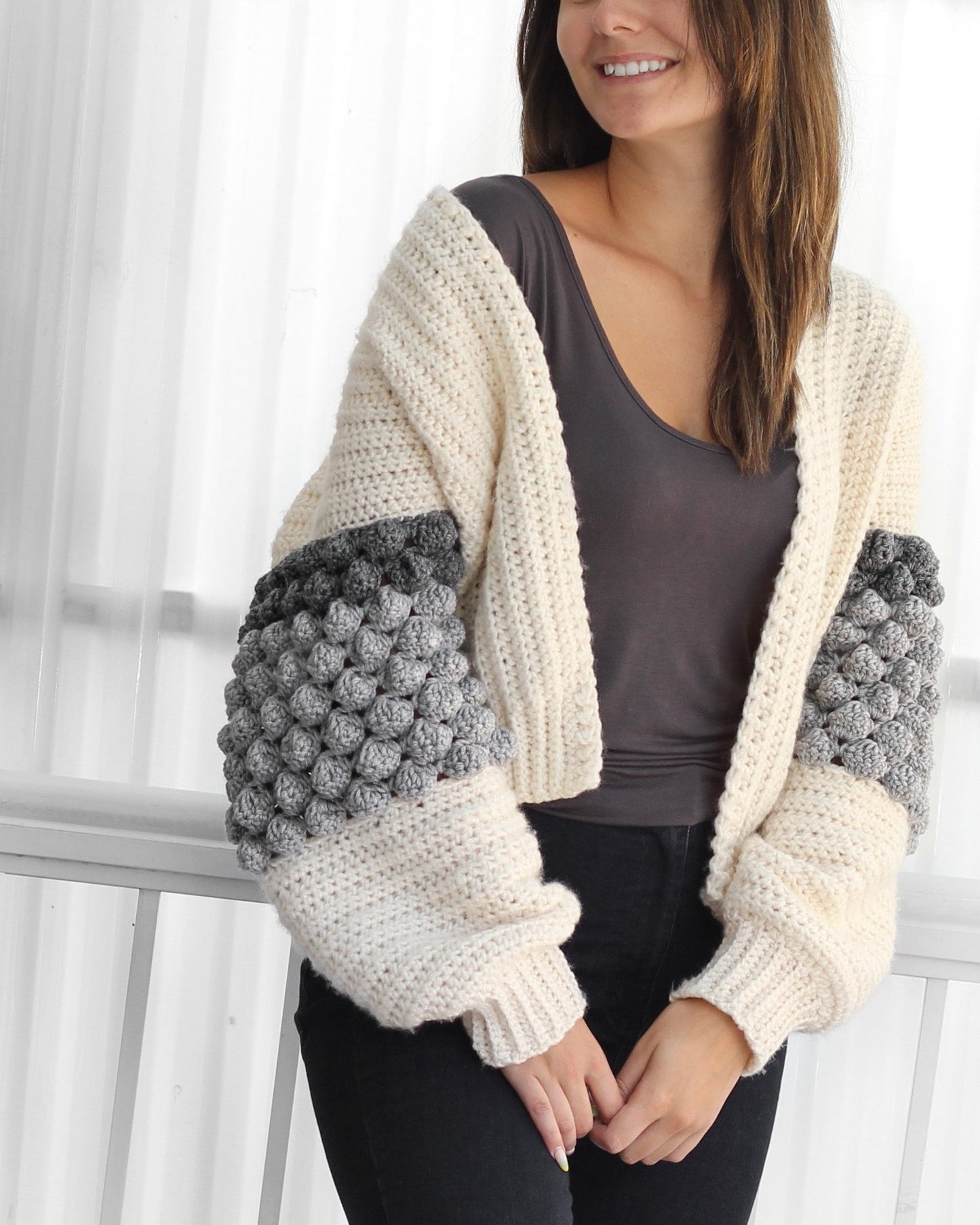 May include: A cream-colored crocheted cardigan with gray and white bobble stitch detailing on the sleeves. The cardigan has a relaxed fit and a slightly cropped length.