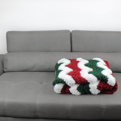 May include: A white, green, and red crocheted throw blanket with a wavy pattern. The blanket is folded and placed on a gray couch.