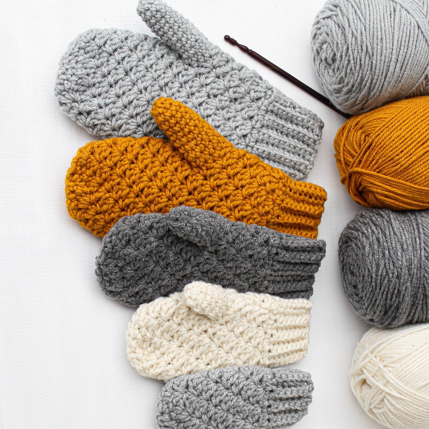 May include: A set of five crocheted mittens in various colors, including gray, mustard yellow, and cream. The mittens are arranged in a row, with the gray mittens at the ends and the mustard yellow and cream mittens in the middle.
