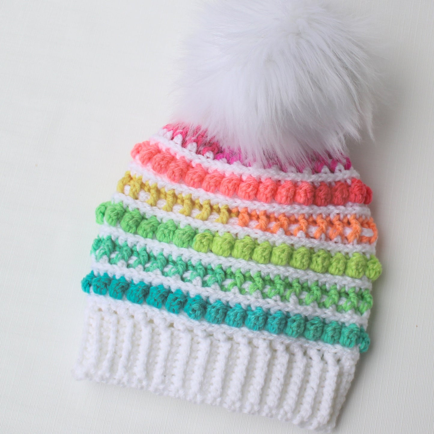 May include: A white crocheted beanie with a rainbow pom-pom trim and a white faux fur pom-pom on top.