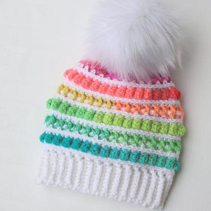 May include: A white crocheted beanie with a rainbow pom-pom trim and a white faux fur pom-pom on top.