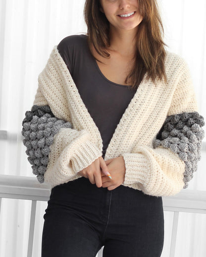 May include: A cream-colored crocheted cardigan with grey bobble stitch detailing on the sleeves. The cardigan is open in the front and has a V-neck.