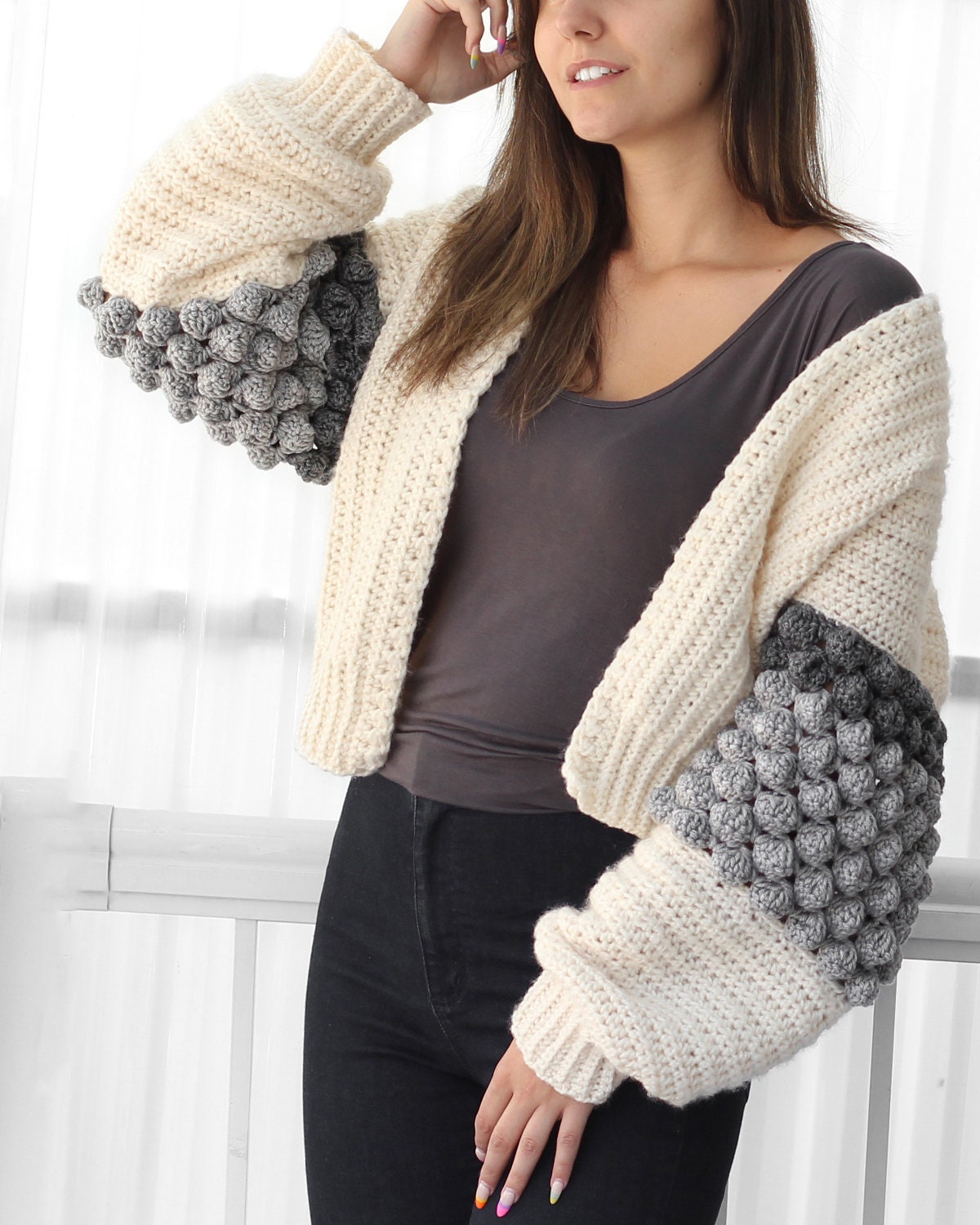 May include: A cropped cardigan with a white, gray, and dark gray color block pattern. The sleeves feature a textured stitch with raised bumps.