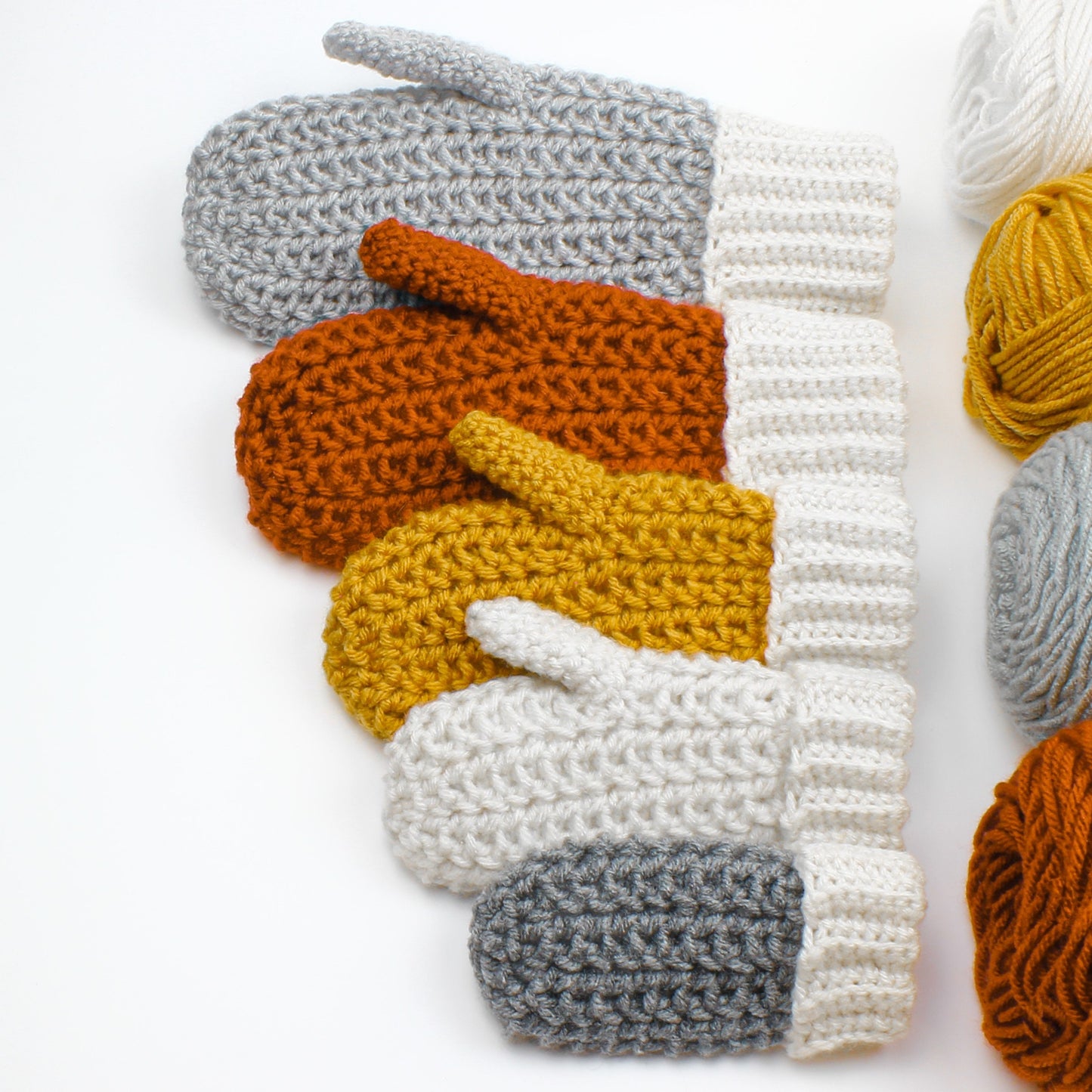 May include: Five crocheted mittens in different colors, including gray, orange, yellow, and white. The mittens are arranged in a row, with the largest mitten at the top and the smallest mitten at the bottom.