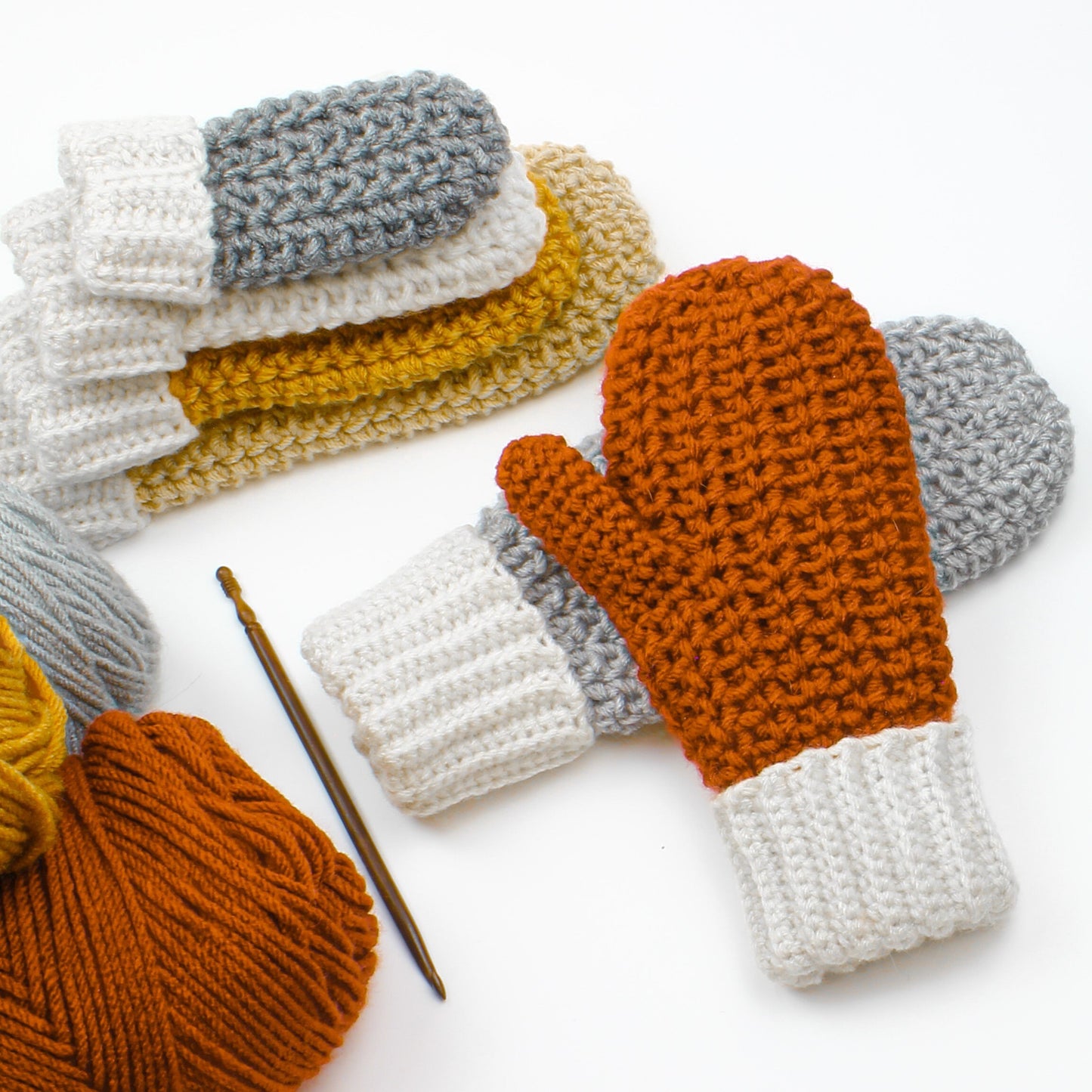 May include: A set of crocheted mittens in various colors, including gray, white, yellow, and orange. The mittens are stacked on top of each other, with a crochet hook lying next to them. There are also balls of yarn in the colors of the mittens.