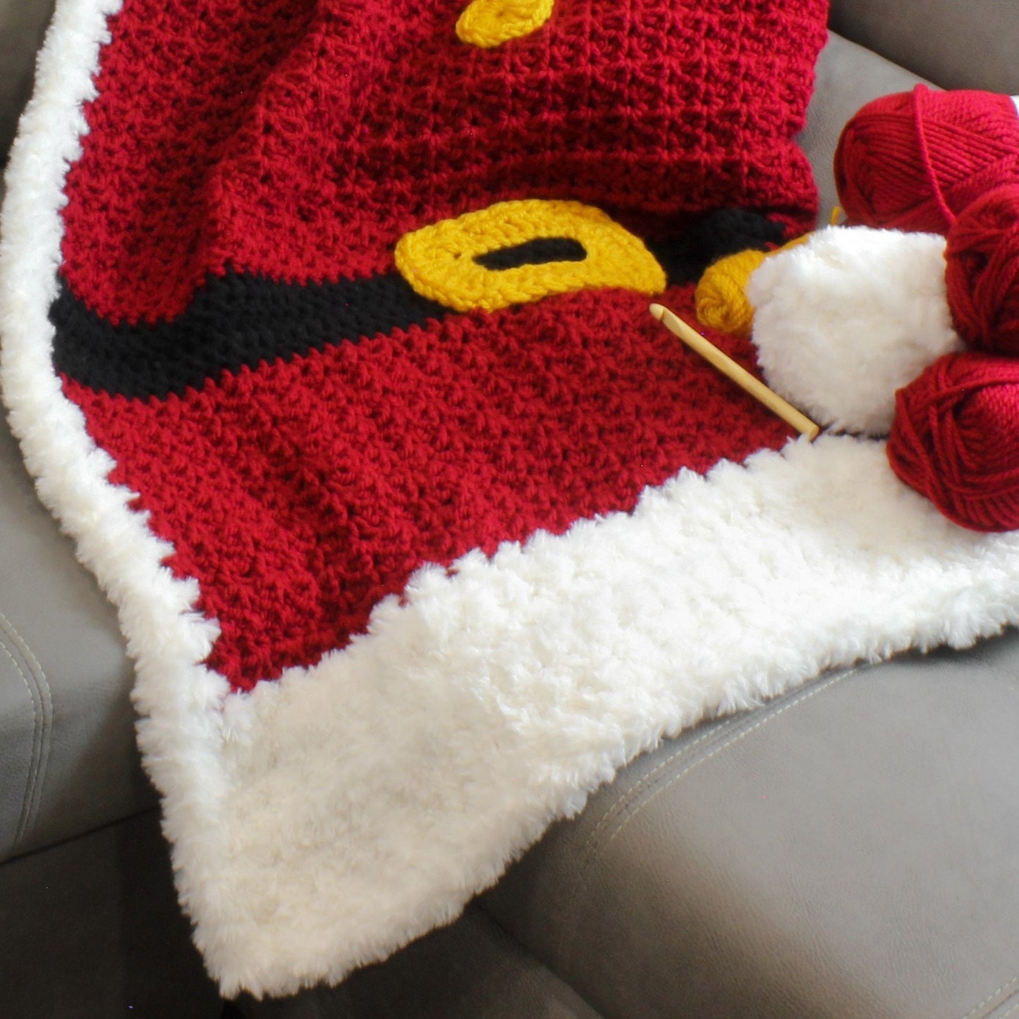 May include: A red, black, and yellow crocheted blanket with a white fluffy trim. The blanket is on a gray surface with yarn balls and a crochet hook.
