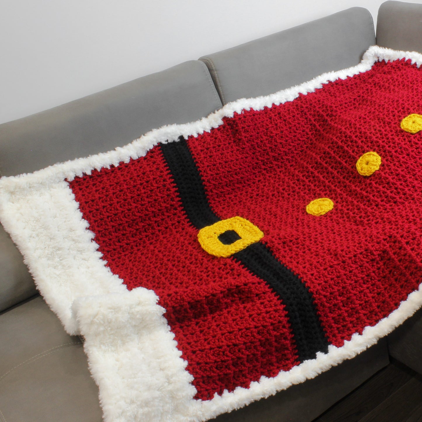 May include: A red crocheted blanket with a black belt and yellow buttons, resembling Santa's suit. The blanket has a white fluffy border.