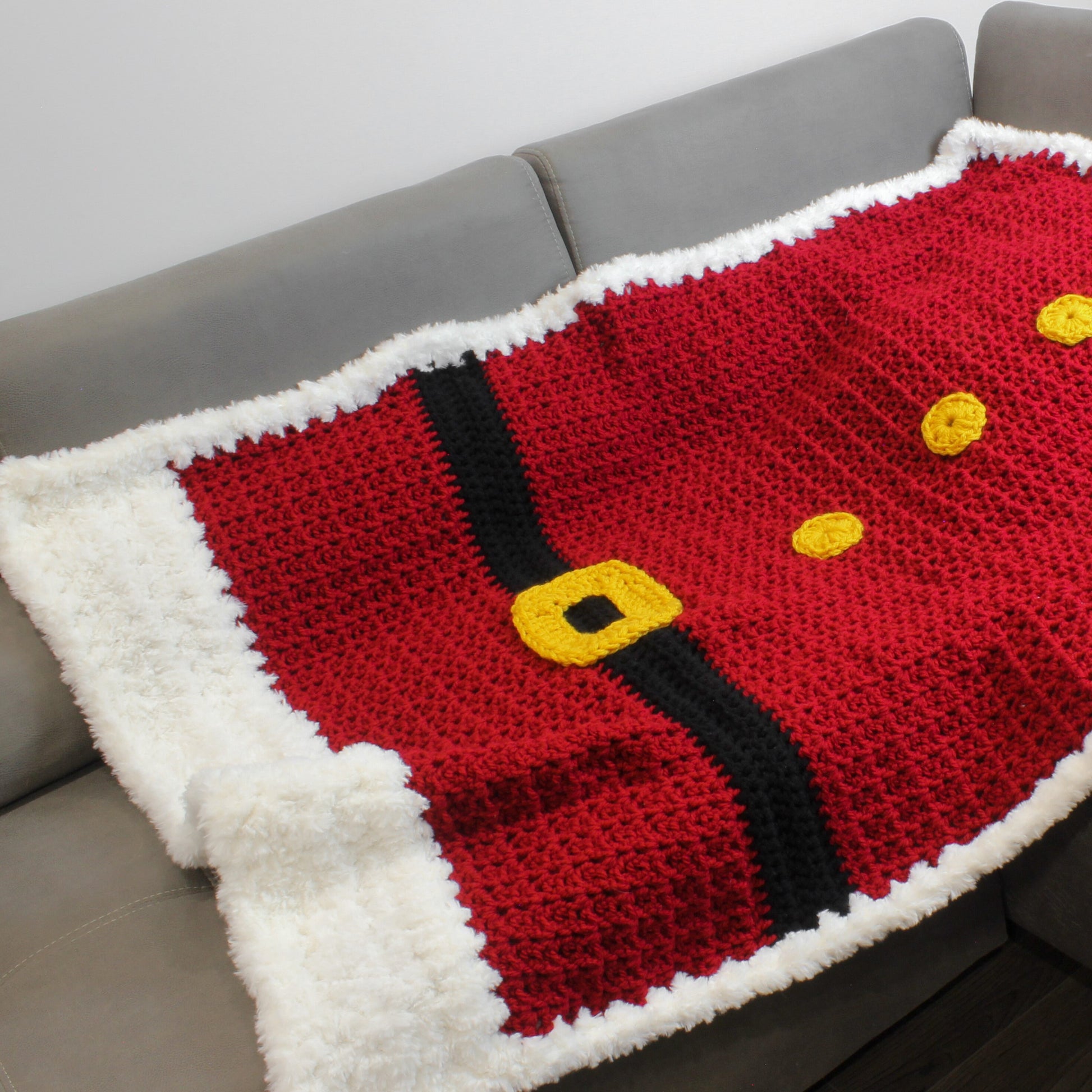 May include: A red crocheted blanket with a black belt and yellow buttons, resembling Santa's suit. The blanket has a white fluffy border.