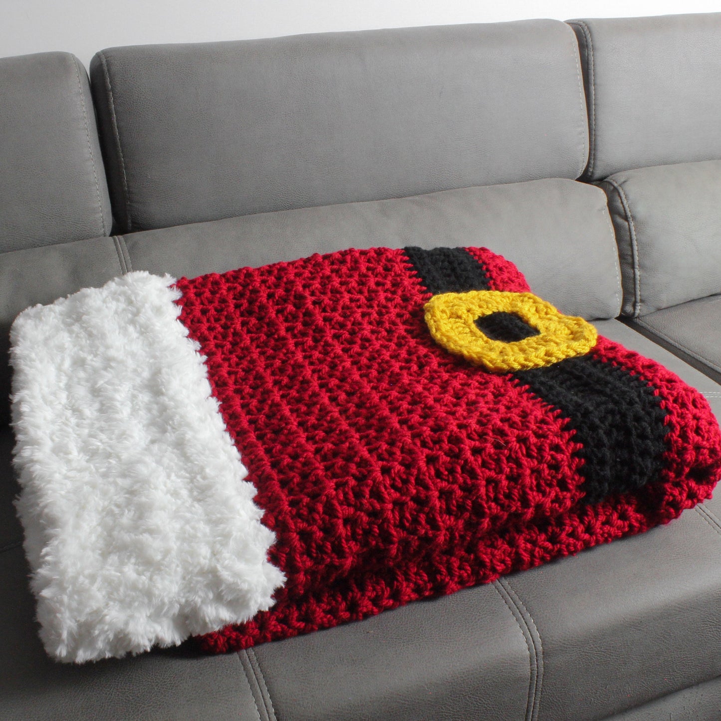 May include: A red, white, and black crocheted blanket with a yellow belt buckle design. The blanket is folded and placed on a gray couch.