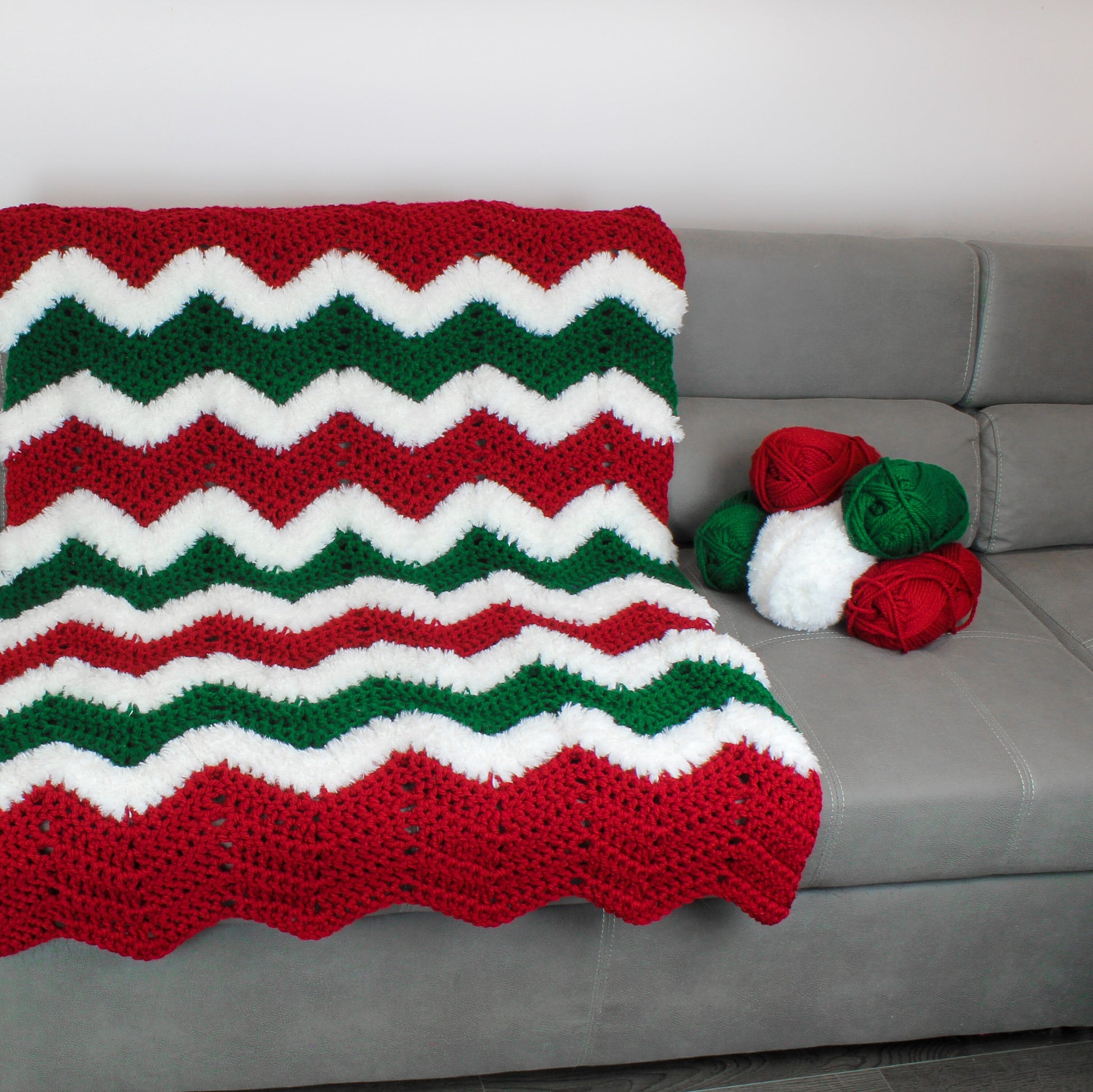 May include: A red, white, and green crocheted blanket with a chevron pattern. The blanket is on a gray couch with balls of yarn in the same colors.