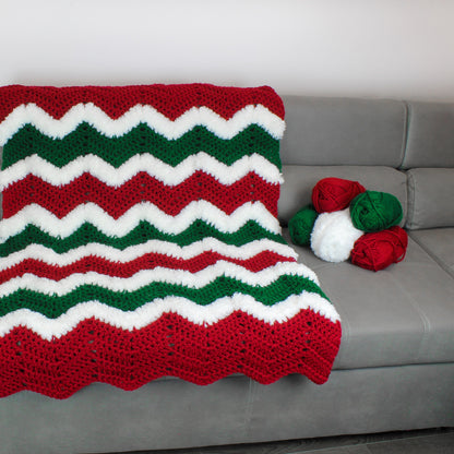 May include: A red, white, and green crocheted blanket with a chevron pattern. The blanket is on a gray couch with balls of yarn in the same colors.