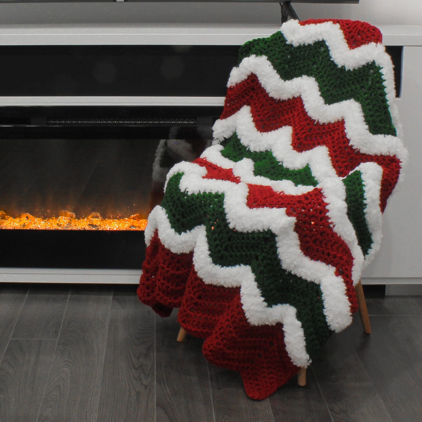 May include: A red, green, and white crocheted blanket with a wavy pattern. The blanket is draped over a chair in front of a fireplace.