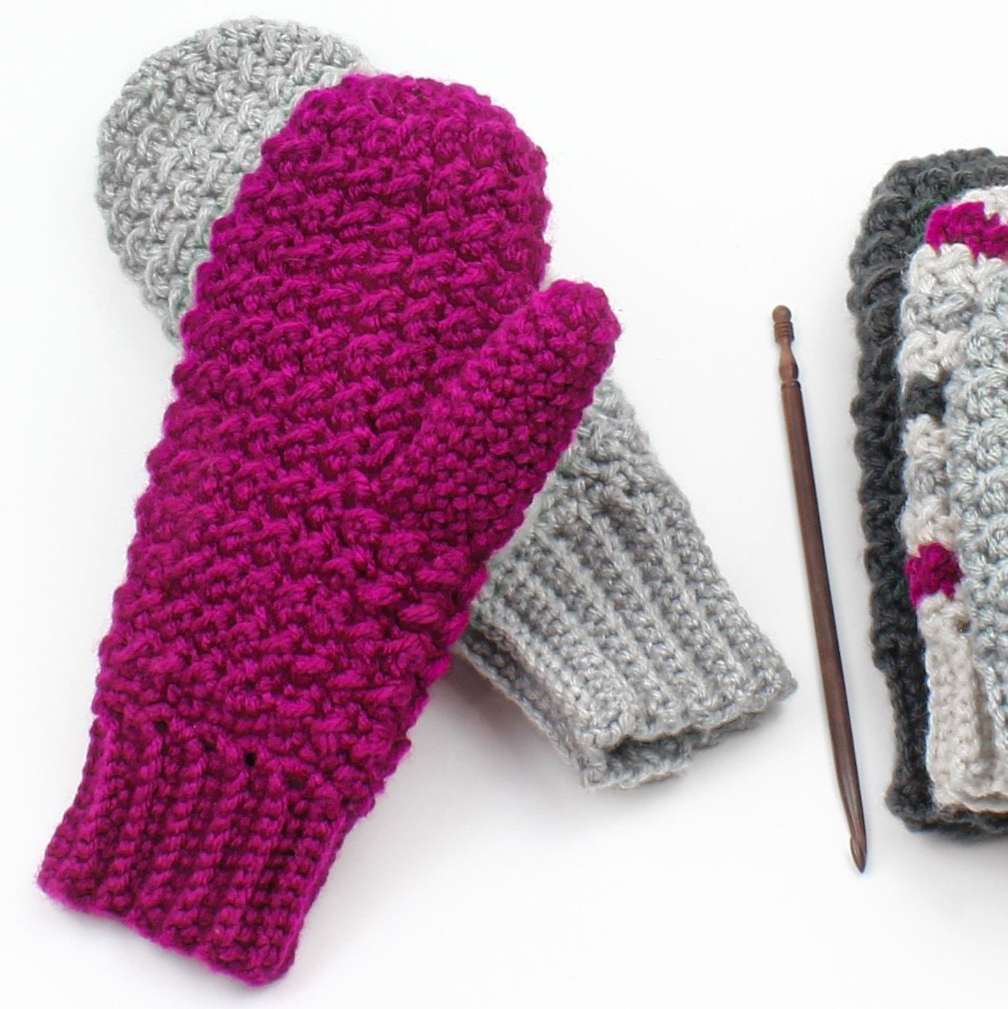 May include: Two pairs of crocheted mittens, one pair is grey and the other is pink. The pink mittens are folded over the grey mittens. A wooden crochet hook is lying on a white surface.
