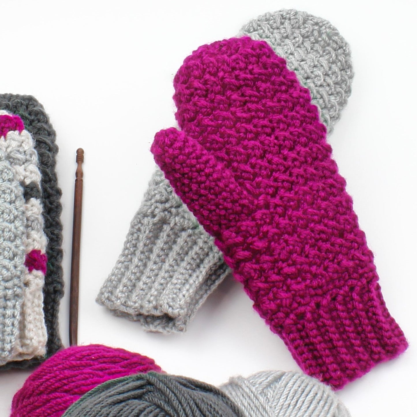 May include: A pair of crocheted mittens, one gray and one pink. The pink mitten has a textured stitch pattern. The mittens are lying on a white surface with yarn balls in the foreground.