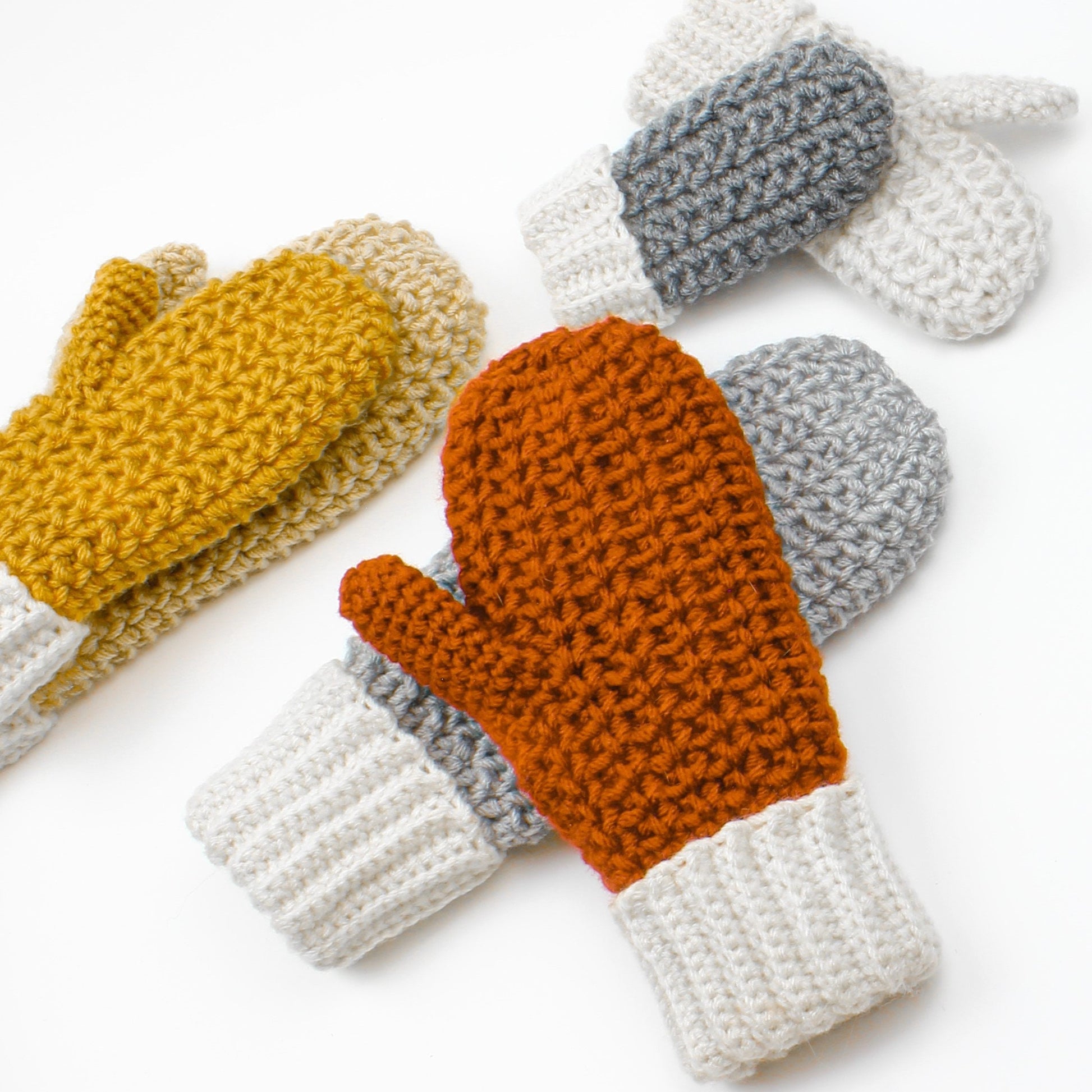 May include: Three pairs of crocheted mittens in different colors. One pair is mustard yellow, one pair is orange, and one pair is gray.
