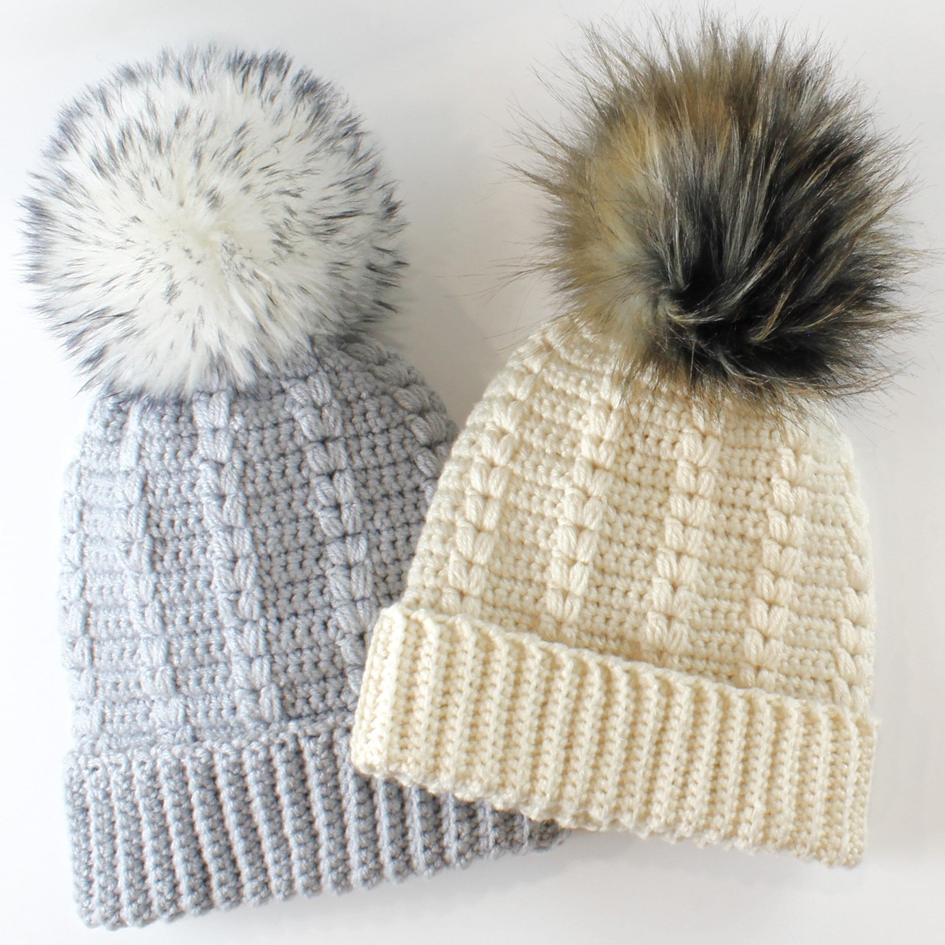 May include: Two crocheted winter hats with faux fur pom poms. One hat is gray and the other is cream.