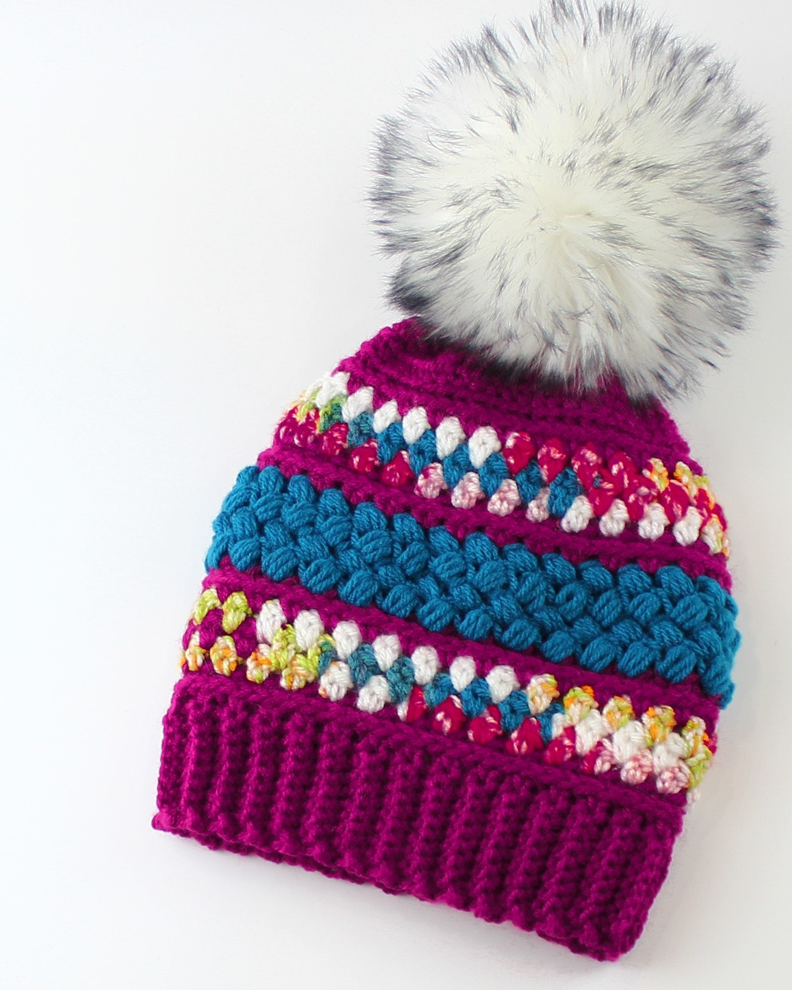 May include: A pink crocheted beanie with a white faux fur pom pom. The beanie has colorful stripes of blue, white, and pink yarn.