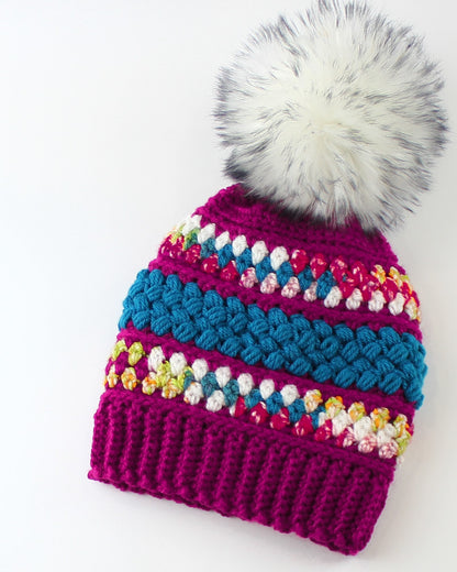 May include: A pink crocheted beanie with a white faux fur pom pom. The beanie has colorful stripes of blue, white, and pink yarn.