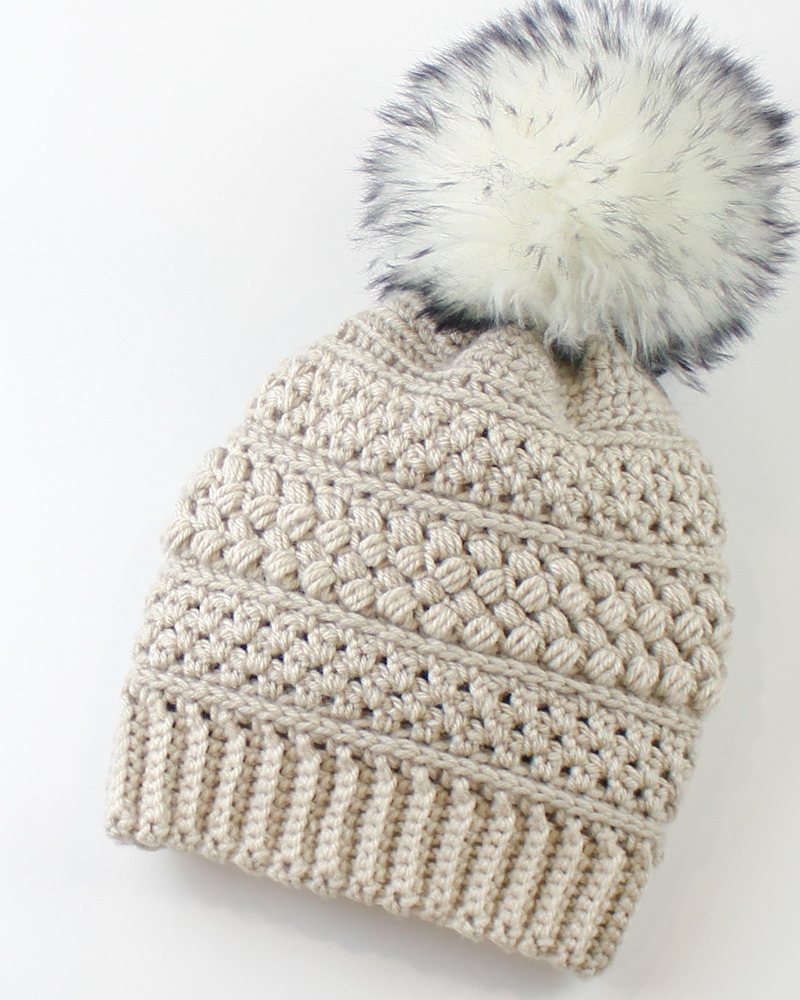 May include: A beige crocheted beanie with a white faux fur pom pom. The beanie has a textured stitch pattern.