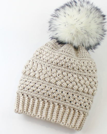 May include: A beige crocheted beanie with a white faux fur pom pom. The beanie has a textured stitch pattern.