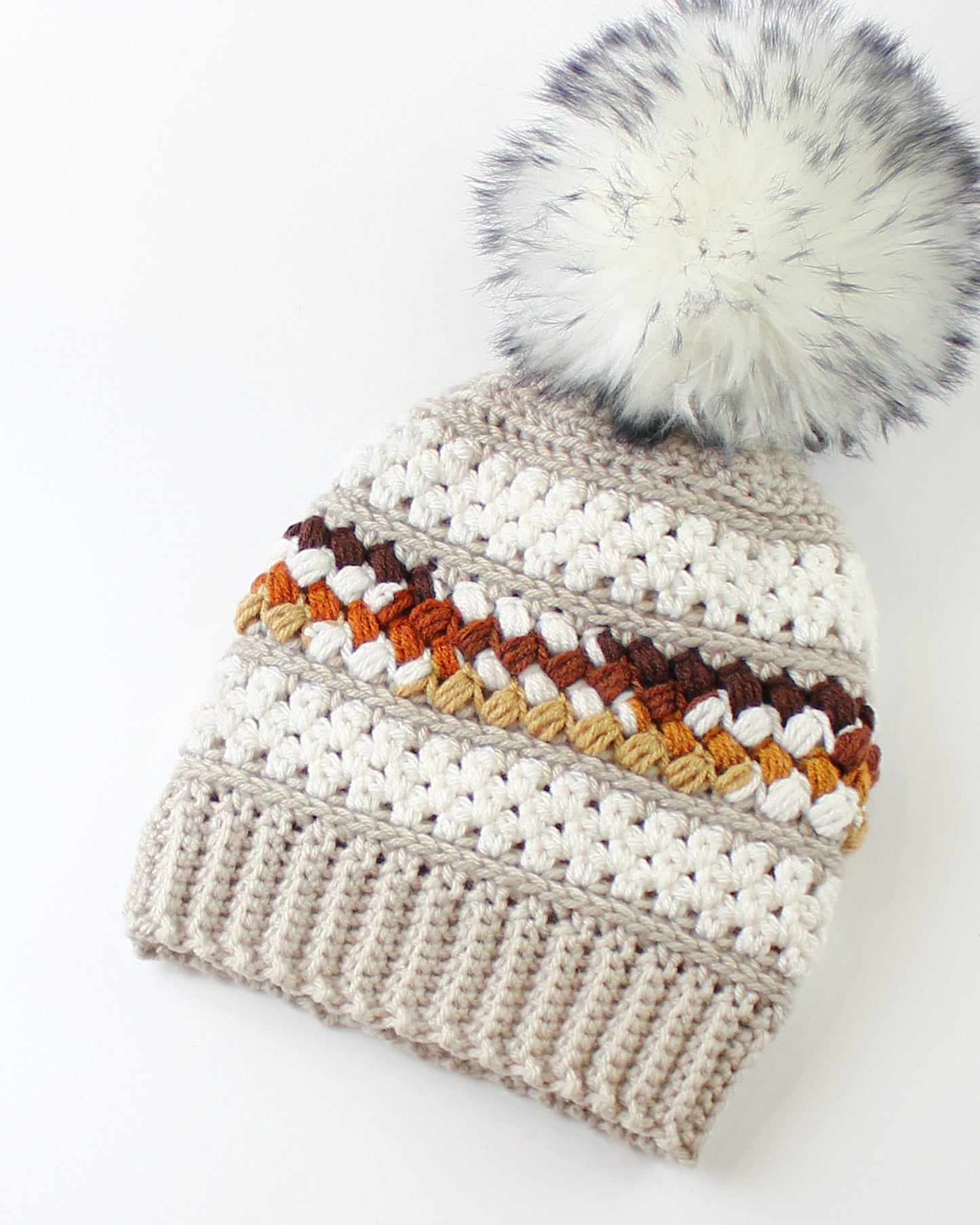 May include: A beige crocheted beanie with a white faux fur pom pom. The beanie has a striped pattern with brown, orange, and beige yarn.