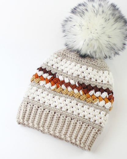 May include: A beige crocheted beanie with a white faux fur pom pom. The beanie has a striped pattern with brown, orange, and beige yarn.