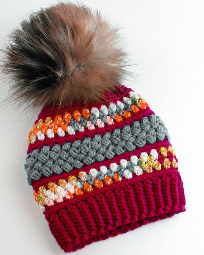 May include: A crocheted beanie with a colorful striped pattern in shades of red, orange, white, and gray. The beanie has a fluffy brown and black pom pom on top.