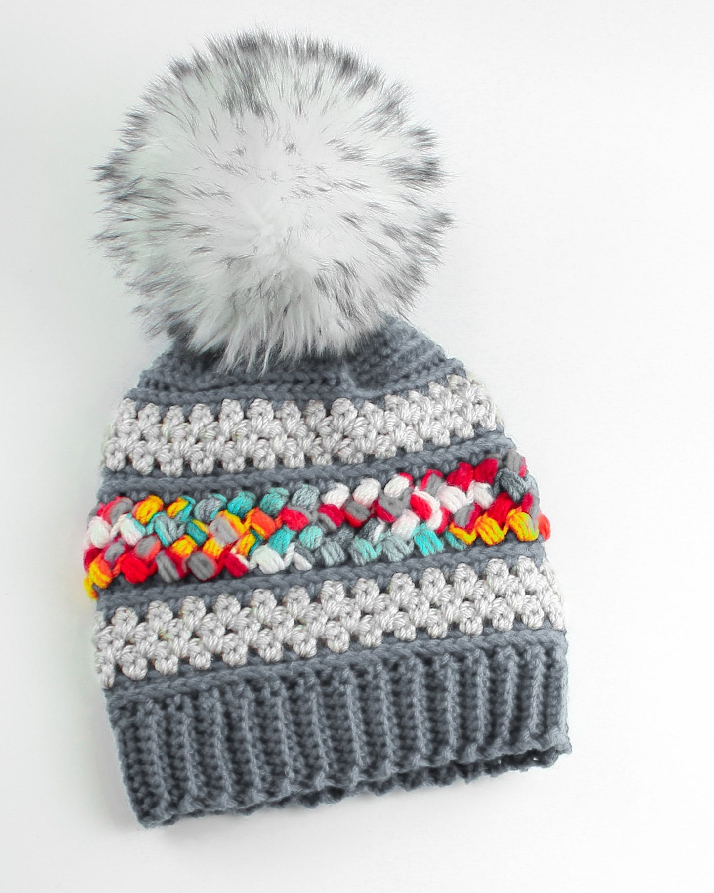 May include: Gray crocheted beanie with a white faux fur pom pom and a colorful braided stripe.