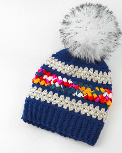 May include: A blue crocheted beanie with a white pom pom. The beanie has a colorful stripe of crocheted yarn in shades of red, orange, yellow, green, blue, and pink.