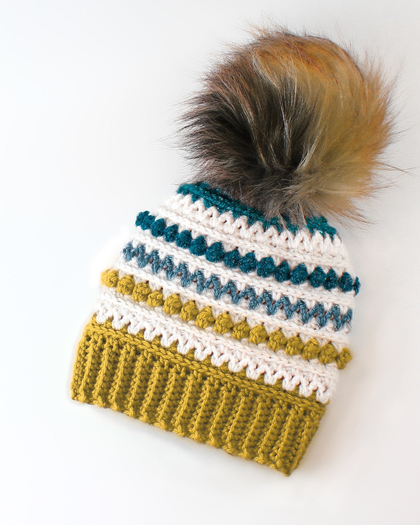 May include: A crocheted beanie with a white, gray, and teal striped pattern and a yellow brim. The beanie has a fluffy brown and beige pompom on top.