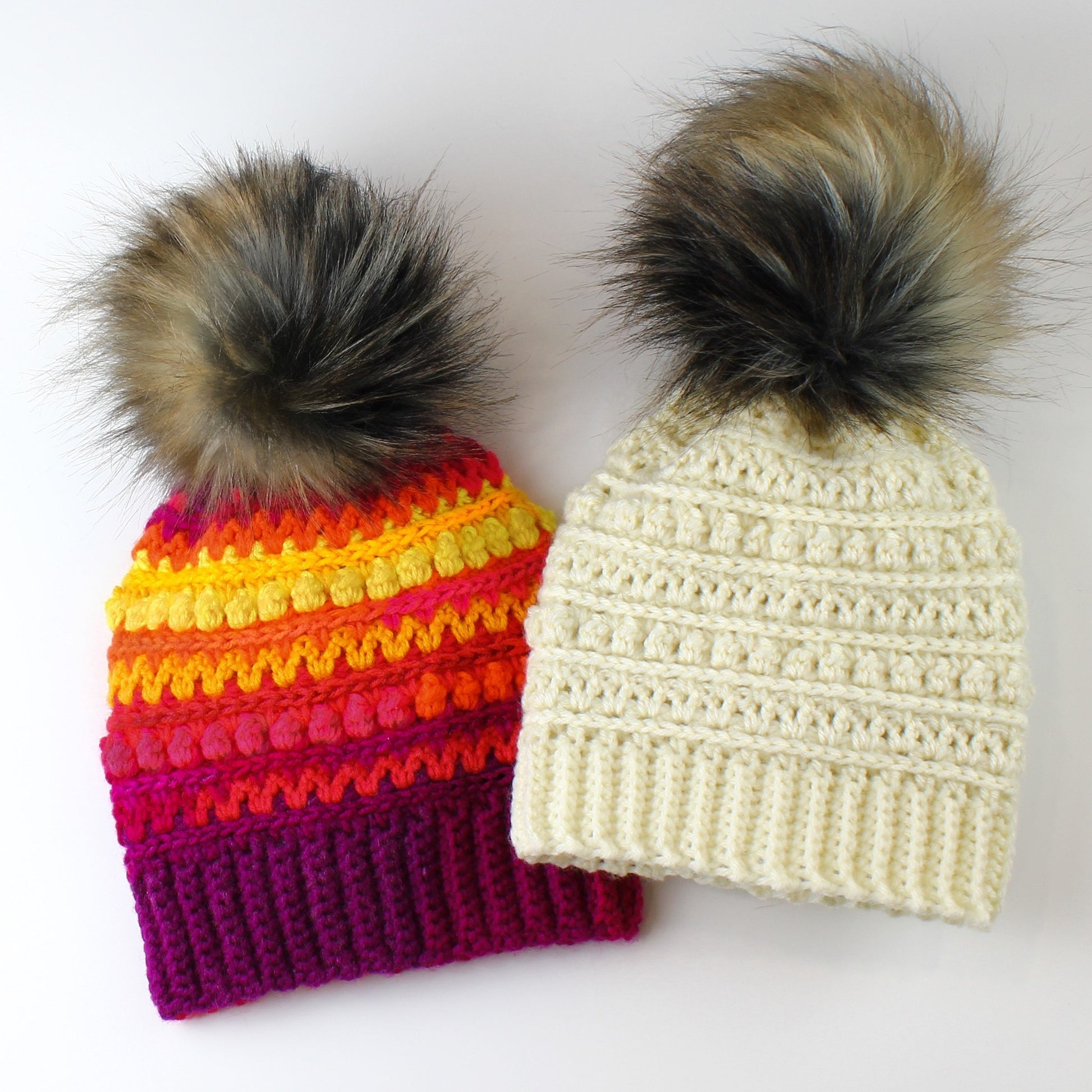 May include: Two crocheted hats with faux fur pom poms. One hat is white with a simple stitch pattern. The other hat has a colorful striped pattern with a pink base.