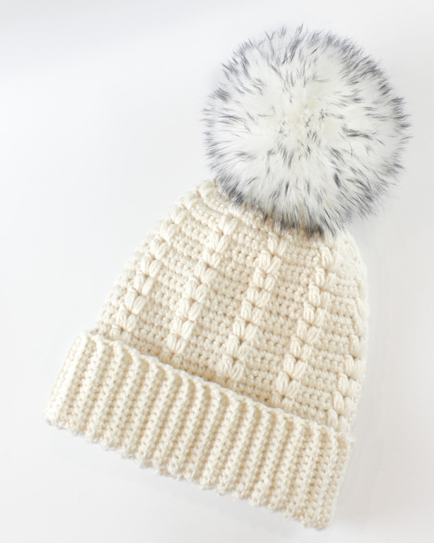 May include: A white crocheted beanie with a fluffy white pom pom. The beanie has a ribbed brim and a textured stitch pattern.