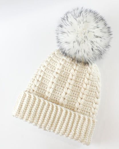 May include: A white crocheted beanie with a fluffy white pom pom. The beanie has a ribbed brim and a textured stitch pattern.