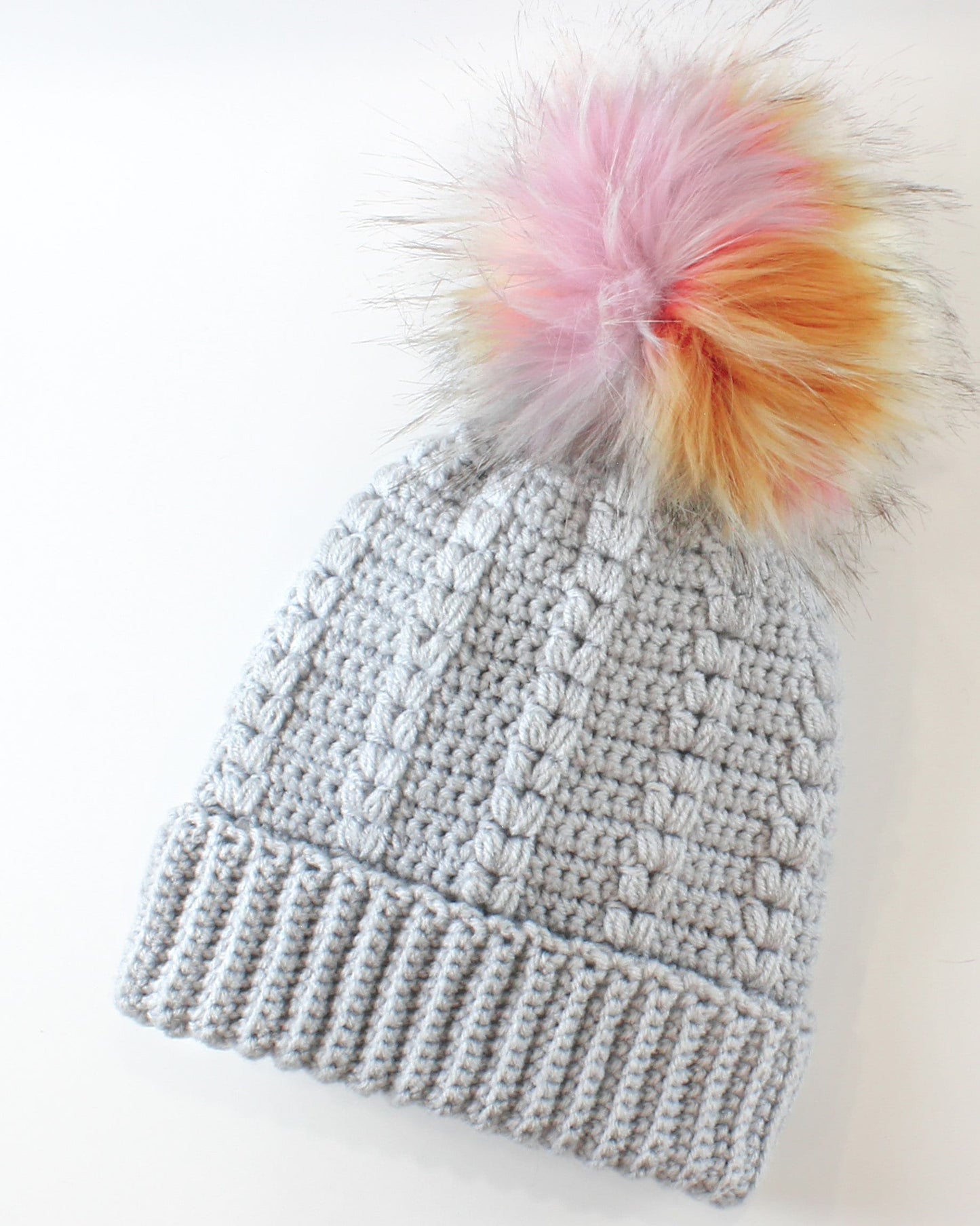 May include: A gray crocheted beanie with a fluffy pom pom in shades of pink, orange, and yellow.