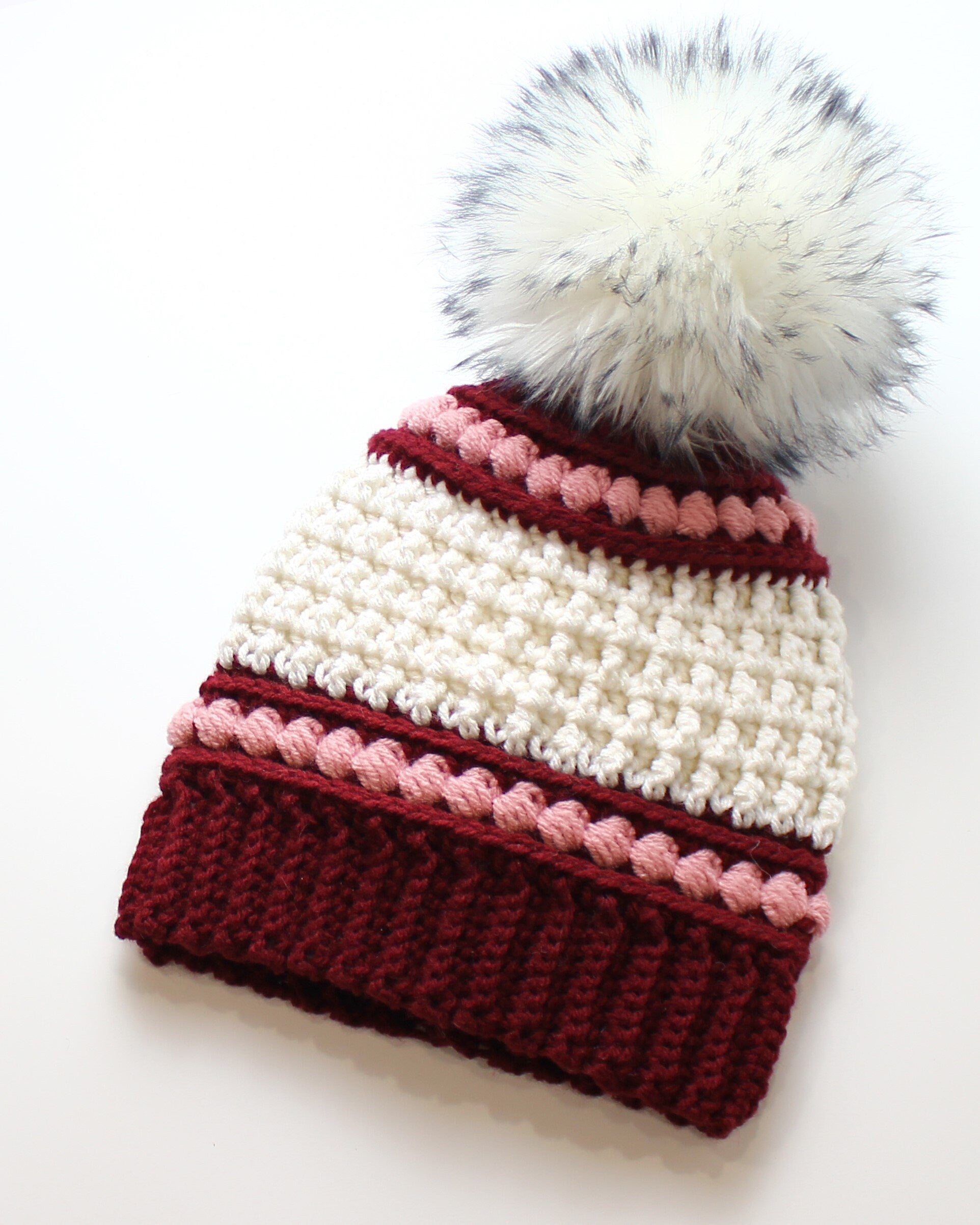 May include: A handmade crochet beanie in burgundy, cream, and pink. The hat features a textured stitch pattern with a ribbed brim and a decorative pink trim. A large, fluffy white and gray faux fur pom-pom tops the hat.