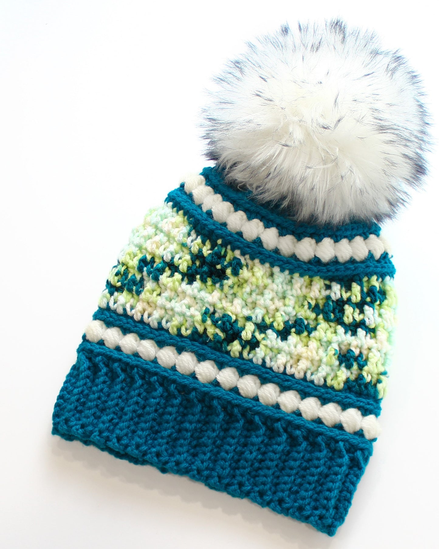 May include: A handmade teal and white knit hat with a large, fluffy white pom-pom. The hat features a textured stitch pattern with white bobble trim and a speckled green and white design. The hat measures approximately 9 inches in height.