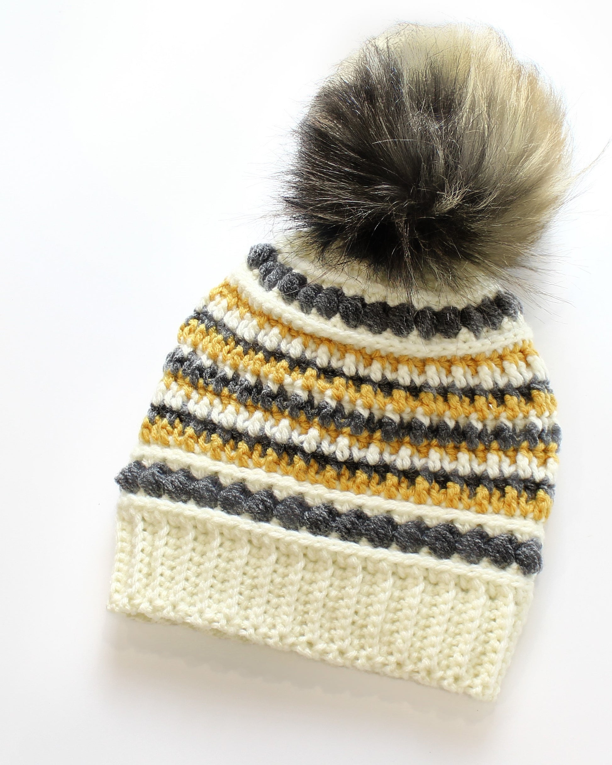 May include: A handmade crochet hat with a faux fur pom-pom. The hat features a cream-colored ribbed brim, with alternating stripes of yellow, white, and gray. The pom-pom is a mix of gray, brown, and cream-colored faux fur.