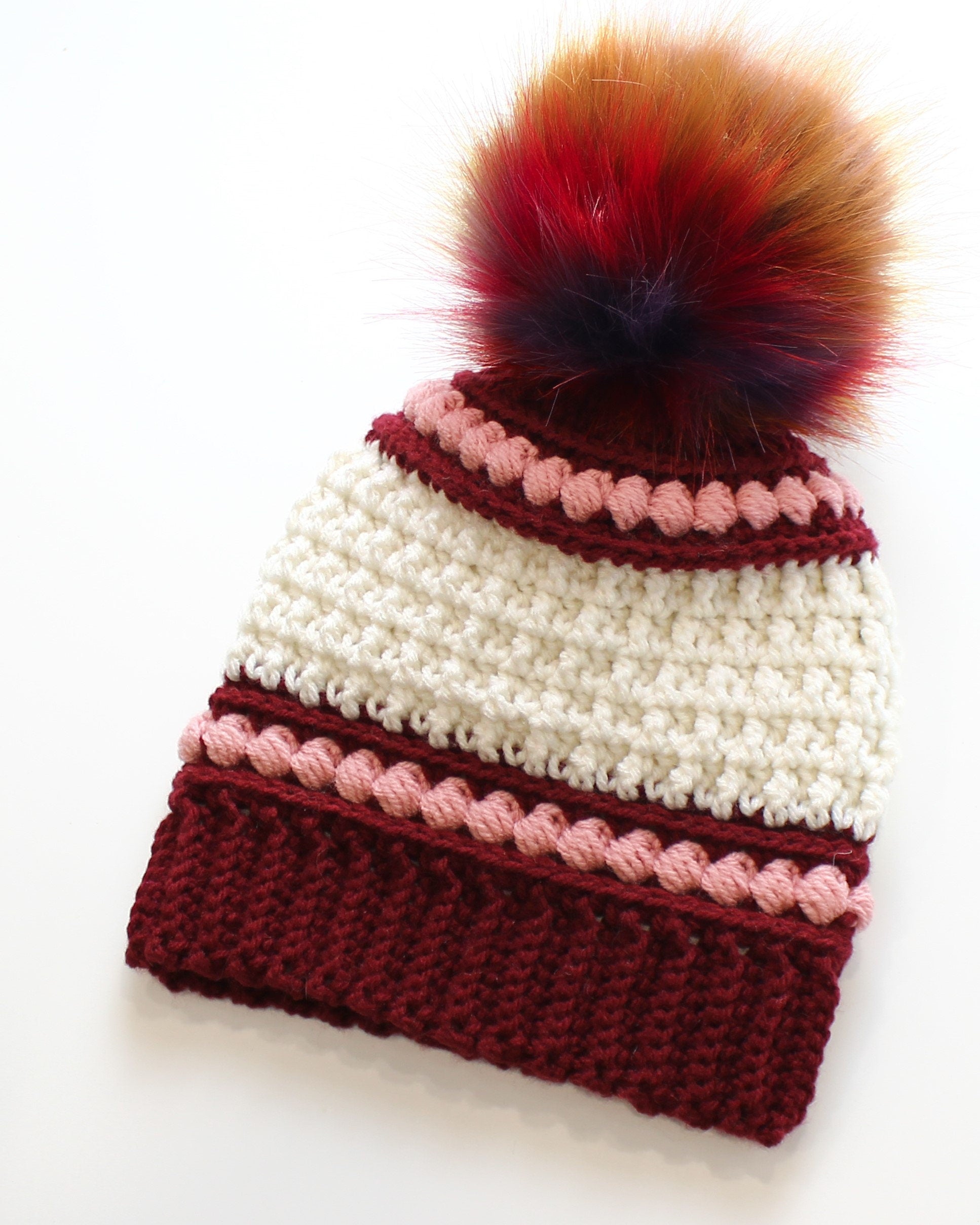 May include: A handmade crochet beanie in cream and burgundy, adorned with a multi-colored faux fur pom-pom. The hat features a ribbed burgundy brim and is accented with pink bobble stitch detailing. A cozy winter accessory.