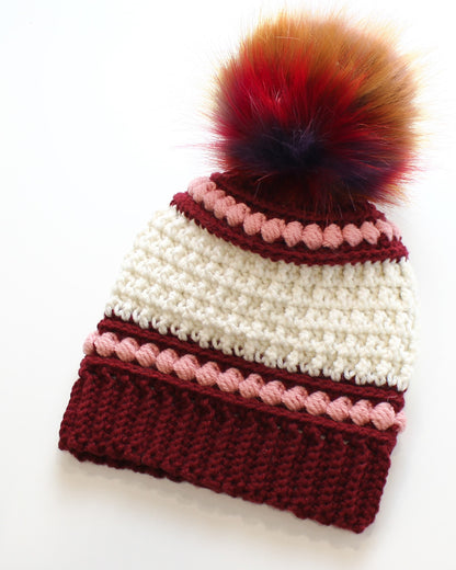 May include: A handmade crochet beanie in cream and burgundy, adorned with a multi-colored faux fur pom-pom. The hat features a ribbed burgundy brim and is accented with pink bobble stitch detailing. A cozy winter accessory.