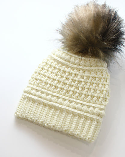 May include: Cream-colored crochet beanie with a large, fluffy, brown and black faux fur pom-pom. The hat features a ribbed brim, a textured body, and a decorative band of bobble stitches. A cozy winter accessory.