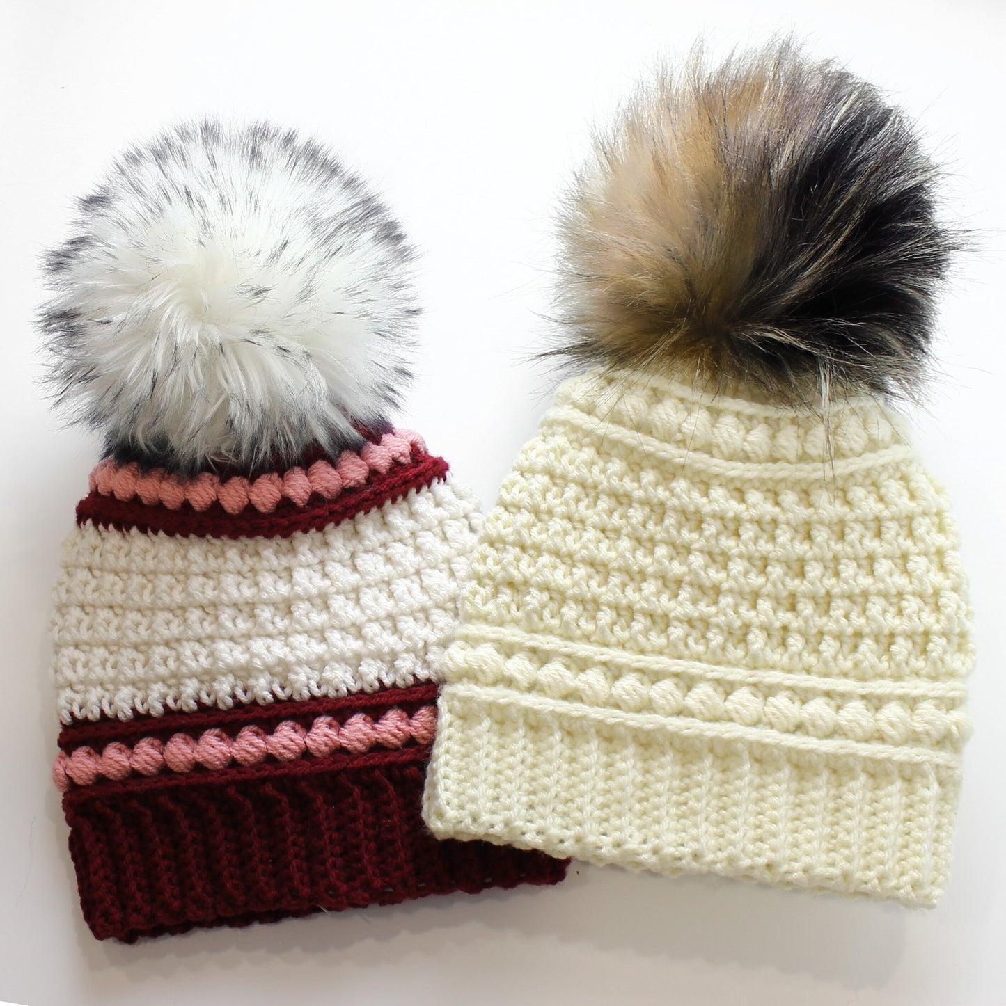 May include: Two handmade crochet beanies with faux fur pom-poms. One beanie is cream-colored with a brown and black pom-pom. The other beanie is white with a burgundy, pink, and white striped design and a white and gray pom-pom.